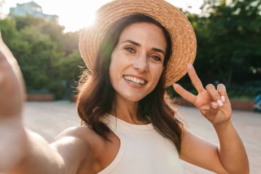 Hasır şapka takan, selfie çeken ve yaz parkında yürürken barış işareti yapan orta yaşlı tatlı bir kadın.