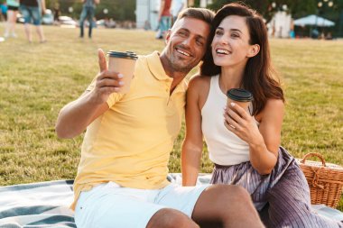 Heyecanlı orta yaşlı bir çiftin portresi. Kahve içip parktaki çimlerde otururken işaret eden kadın ve erkek.