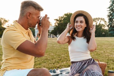 Yaz parkında çimlerin üzerinde otururken gülümseyen bir kadının fotoğrafını çeken orta yaşlı yakışıklı bir adam.