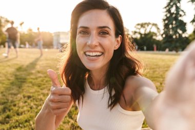 Orta yaşlı neşeli bir kadın resmi. Başparmağını kaldırıyor ve yaz parkında çimlerin üzerinde otururken selfie çekiyor.