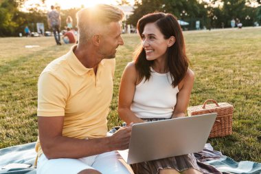 Yaz parkında piknik yaparken dizüstü bilgisayar kullanarak gülümseyen orta yaşlı bir çiftin portresi.