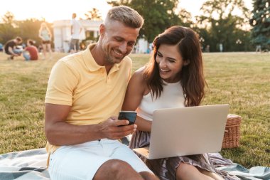 Yaz parkında piknik yaparken dizüstü bilgisayar kullanan gülümseyen orta yaşlı çift ve kadının portresi.