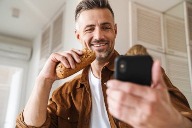 Beyaz saçlı, neşeli bir adamın akıllı telefonuyla selfie çekerken beyaz mutfakta öğle yemeği yediği görüntüsü.