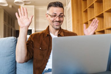 Neşeli yetişkin bir adamın evde kanepede otururken el sallaması ve dizüstü bilgisayarla çalışması.