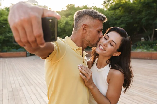 幸せな大人のカップルの男と女の肖像写真を携帯電話で撮影し 夏の公園を歩きながらキス — ストック写真