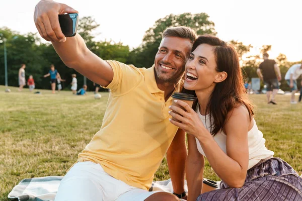 一对快乐的成年夫妇坐在公园的草地上 一边喝着咖啡一边用手机拍照的画像 — 图库照片