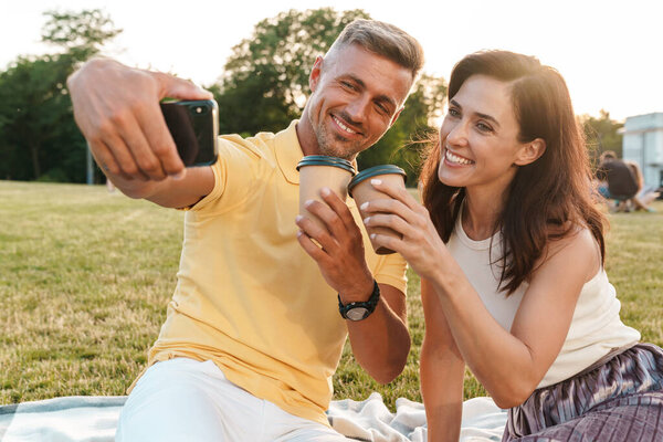 Portrait of smiling adult couple man and woman drinking coffee takeaway and taking selfie photo on cellphone while sitting on grass in park