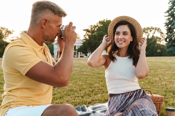 Yaz parkında çimlerin üzerinde otururken gülümseyen bir kadının fotoğrafını çeken orta yaşlı yakışıklı bir adam.