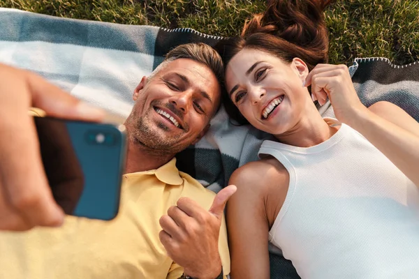 一对积极的成年恋人躺在一个美丽的绿色自然公园里 用手机自拍 竖起大拇指 — 图库照片