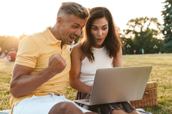 Portrait of surprised middle-aged couple man and woman screaming and celebrating success while using laptop computer in summer park