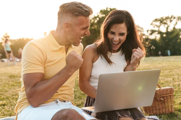 Portrait of excited middle-aged couple man and woman screaming and celebrating success while using laptop computer in summer park