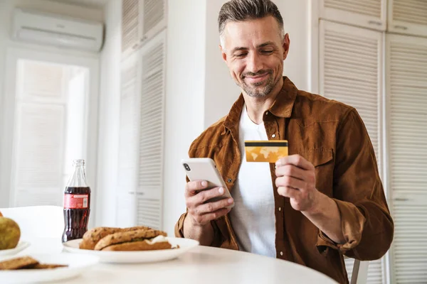 Image Handsome Joyful Man Holding Credit Card Using Smartphone While — Stock Photo, Image