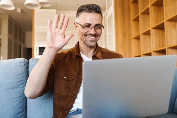 Kép Vidám Felnőtt Férfi Integetett Kezét Dolgozik Laptop Miközben Kanapén — Stock Fotó