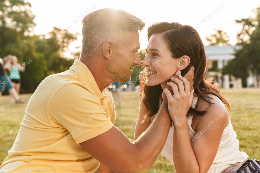 Portrait of lovely middle-aged couple man and woman looking at each other and hugging while sitting on grass in summer park