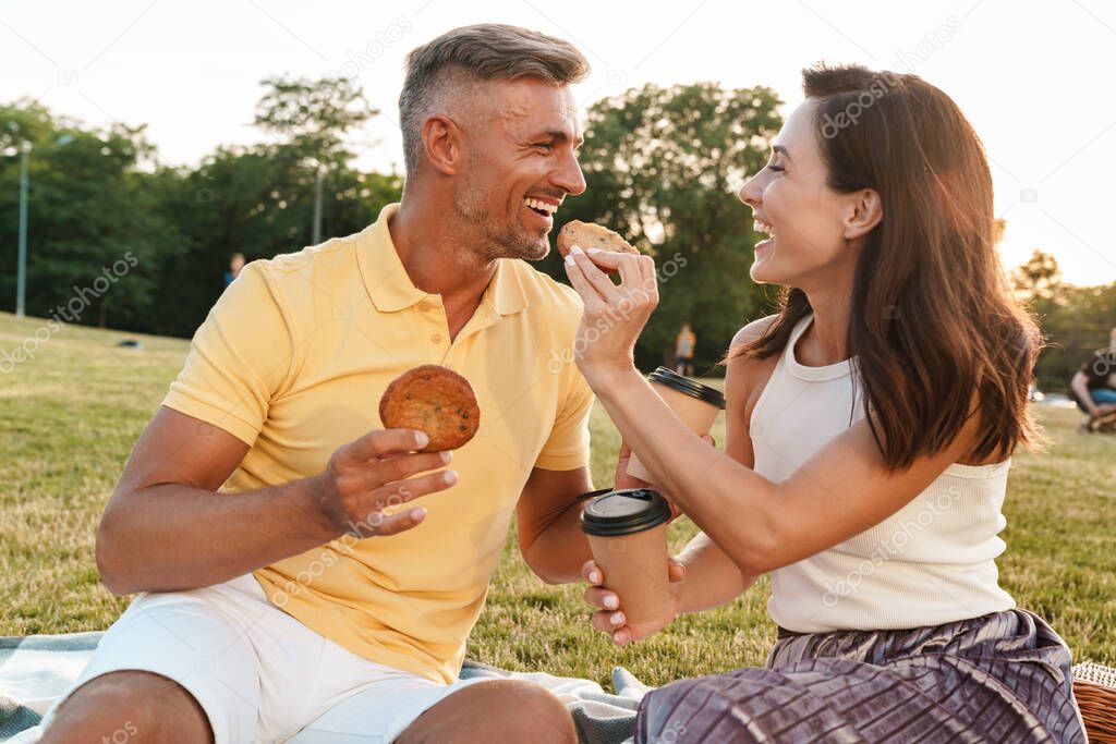 Portrait of amazed middle-aged couple man and woman drinking coffee takeaway and eating cookies while sitting on grass in park