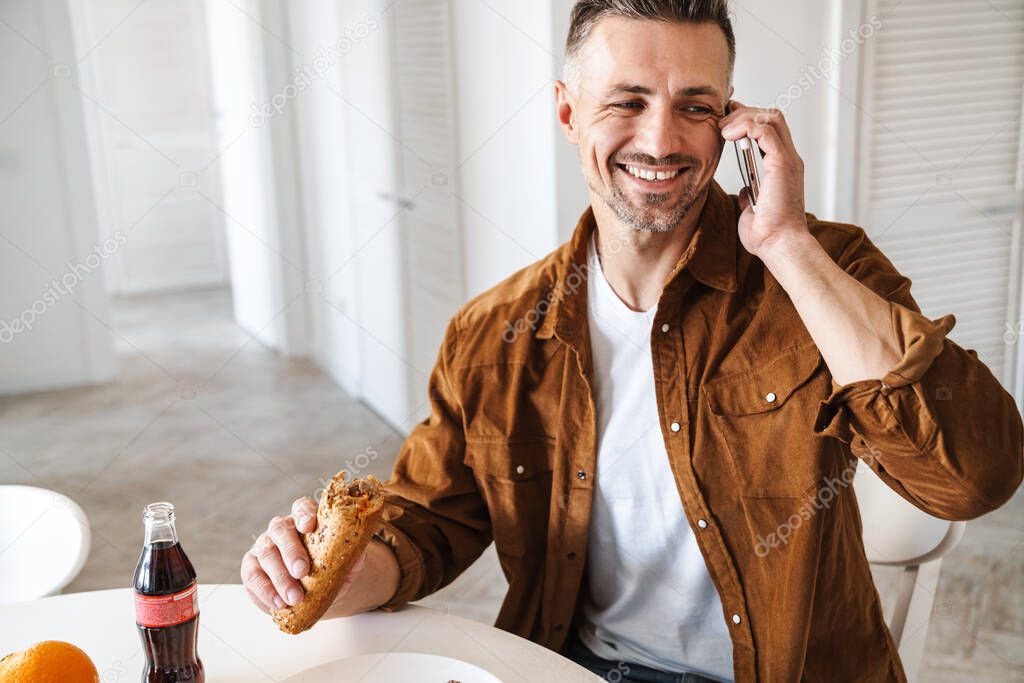 Image of grey-haired joyful man smiling and talking smartphone while have lunch in white kitchen