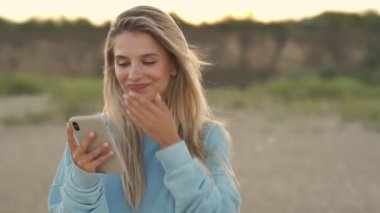 Gülümseyen sarışın bir kız kumsalda yürürken akıllı telefonunu kullanıyor.