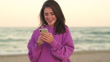 Pozitif bir kız sahilde yürürken akıllı telefonunu kullanıyor.