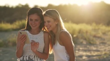 İki güzel kız sahilde yürürken akıllı telefondan fotoğraflara bakıyor.