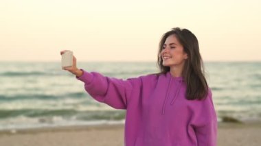 Mutlu bir kız sahilde yürürken kendi fotoğrafını çekerken akıllı telefonunu kullanıyor.