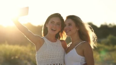 İki pozitif kız sahilde yürürken akıllı telefondan selfie çekiyorlar.