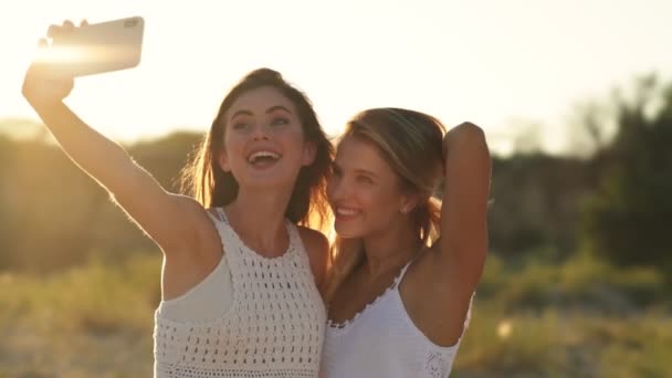 2人の笑顔の女の子がビーチを歩くスマートフォンで写真を撮っています — ストック動画