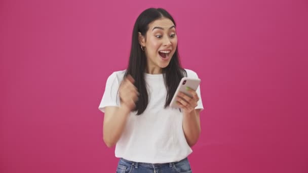 一位惊讶的女士正凝视着她的智能手机 在粉色背景下孤立无援地做着获胜的手势 — 图库视频影像