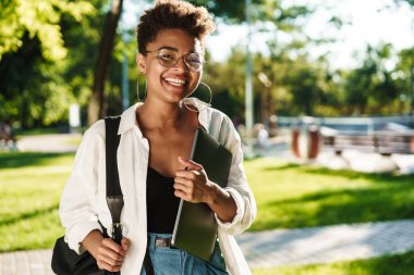 Elinde dizüstü bilgisayarla parkta gezen mutlu bir Afrikalı kadının fotoğrafı.