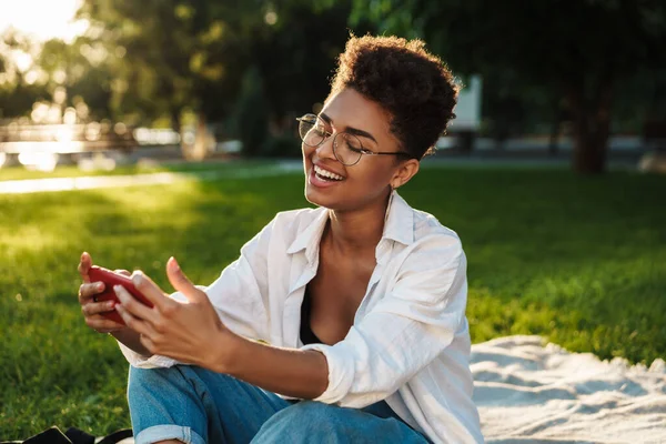 İyimser memnun Afrikalı bir kadının çimlerin üzerinde oturduğu ve cep telefonu kullandığı bir fotoğraf.
