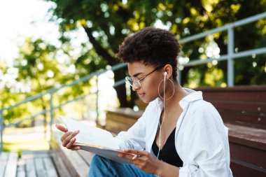 Pozitif yoğunlaştırılmış Afrikalı bir kadının parkta oturmuş müzik dinlerken ve kitap okurken fotoğrafı.