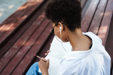 Afro-Afrikalı bir kadının parkta oturmuş müzik dinlerken ve ödev yaparken notlar yazarken fotoğrafı.