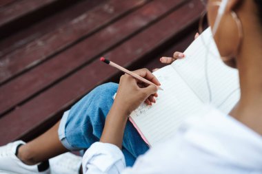 Afro-Afrikalı bir kadının açık havada parkta oturmuş müzik dinlerken ve ödev yaparken notlar yazarken görüntüsü.