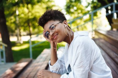 Neşeli Afrikalı pozitifin görüntüsü. Parkta oturmuş kulaklıkla müzik dinliyor.