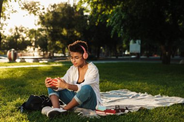 Afrikalı konsantre bir kadın hayal edin. Parkta çimlerin üzerinde oturmuş kulaklıkla müzik dinliyor ve cep telefonu kullanıyor.
