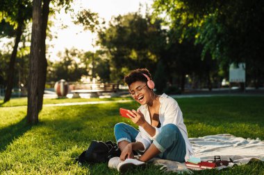 İyimser gülen Afrikalı bir kadının çimlerin üzerinde oturmuş kulaklıkla müzik dinlerken ve cep telefonuyla video izlerken fotoğrafı.