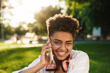 Neşeli bir Afrikalı kadının fotoğrafı. Parkta çimlerin üzerinde oturmuş cep telefonuyla konuşuyor.