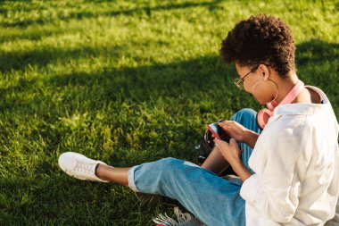 Fotoğrafta gülümseyen iyimser Afrikalı kadın çimlerin üzerinde parkta oturuyor ve cep telefonu kullanıyor.