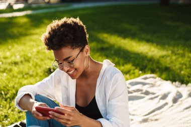 Fotoğrafta gülümseyen iyimser Afrikalı kadın çimlerin üzerinde parkta oturuyor ve cep telefonu kullanıyor.