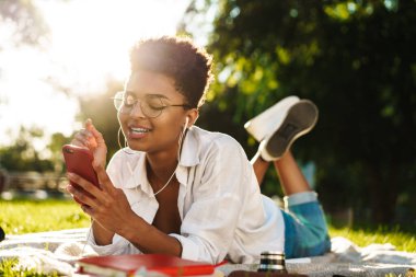 Mutlu, iyimser Afrikalı bir kadının çimlerin üzerinde uzanıp müzik dinlerken cep telefonu kullanmasının fotoğrafı.
