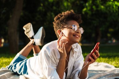 Mutlu gülümseyen Afrikalı kadın portresi çimlerin üzerinde uzanıyor ve müzik dinlerken cep telefonu kullanıyor.
