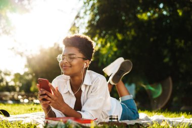 Pozitif Afrikalı bir kadının çimlerin üzerinde dışarıda yatıp müzik dinlerken cep telefonu kullanması.