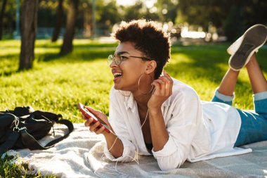 Neşeli Afrikalı bir kadının çimlerin üzerinde uzanıp müzik dinlerken cep telefonu kullanması.