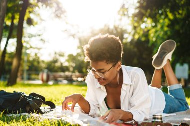 Mutlu, gülümseyen Afrikalı bir kadının çimlerin üzerinde uzanmış kitap okuduğu görüntüsü.