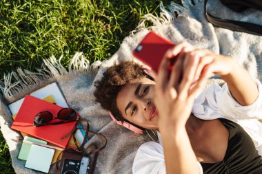 Pozitif Afrikalı bir kadının çimlerin üzerinde uzanmış cep telefonu kullanırken müzik dinlediği görüntüsü