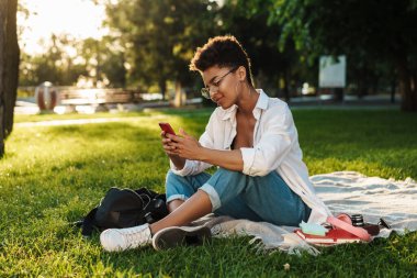 Mutlu Afrikalı bir kadının çimlerin üzerinde parkta oturduğunu ve cep telefonu kullandığını hayal et.