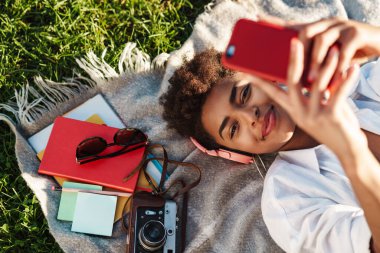 Gülümseyen Afrikalı bir kadının çimlerin üzerinde uzanmış cep telefonuyla müzik dinlediği bir görüntü.