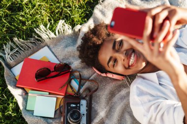 Mutlu, neşeli, Afrikalı bir kadının çimlerin üzerinde uzanıp cep telefonu kullanırken müzik dinlediği görüntüsü.