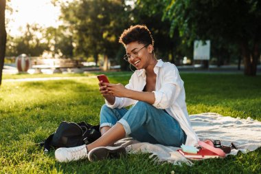 İyimser memnun Afrikalı bir kadının çimlerin üzerinde oturduğu ve cep telefonu kullandığı bir fotoğraf.