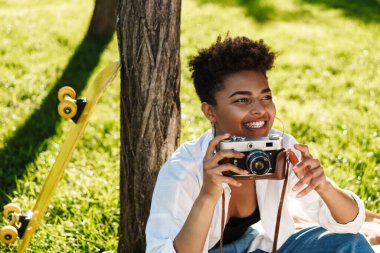Gülümseyen harika bir Afrikalı kadının fotoğrafı. Parkta çimlerin üzerinde oturuyor ve elinde kamera var.