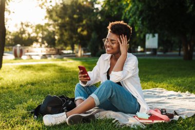 Mutlu Afrikalı bir kadının çimlerin üzerinde parkta oturduğunu ve cep telefonu kullandığını hayal et.
